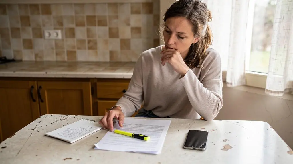Une femme relit attentivement un document papier attablée dans une cuisine lumineuse.